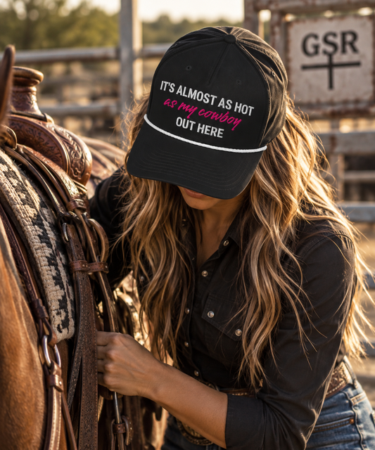 Woman wearing a black Hot As My Cowboy rope cap while saddling a horse at the ranch