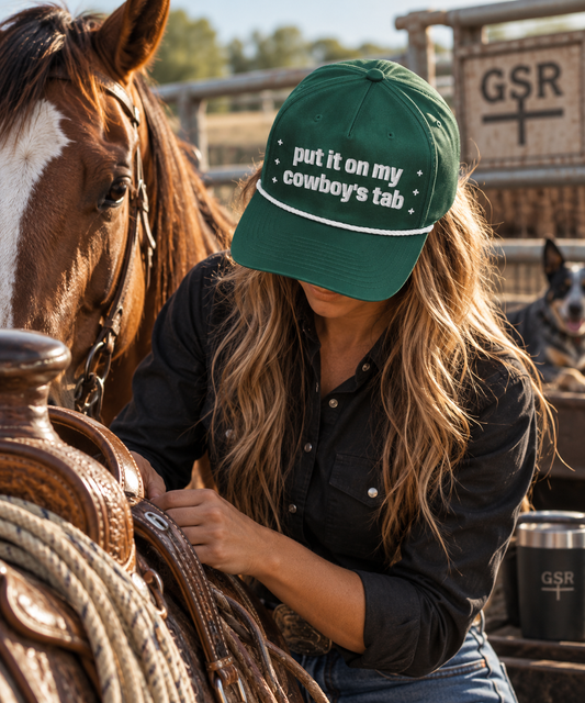 Woman wearing a green Put It on My Cowboy’s Tab rope cap while saddling a horse at the ranch