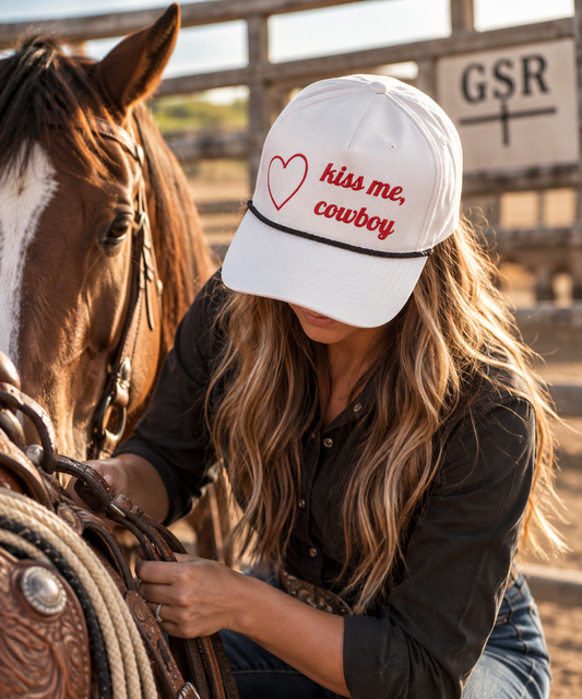 Woman wearing a white Kiss Me Cowboy rope cap while saddling a horse at the ranch