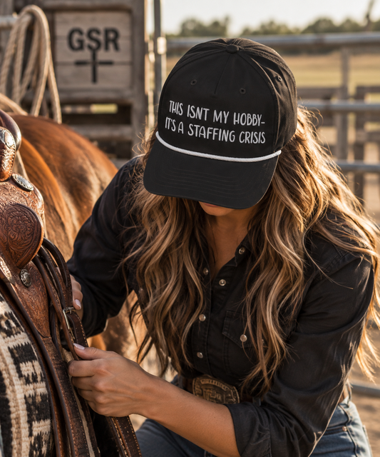 Woman wearing a black Staffing Crisis rope cap while saddling a horse at the ranch